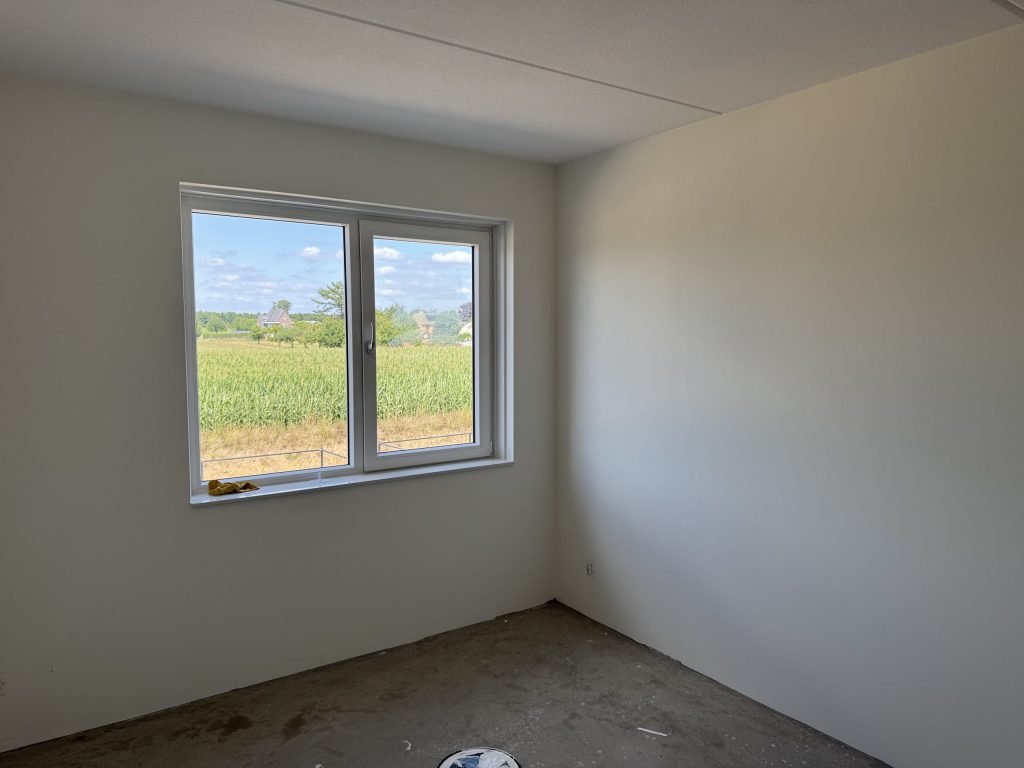 Lege nieuwbouwslaapkamer met gladde witte wanden en een raam dat uitkijkt op een maisveld
